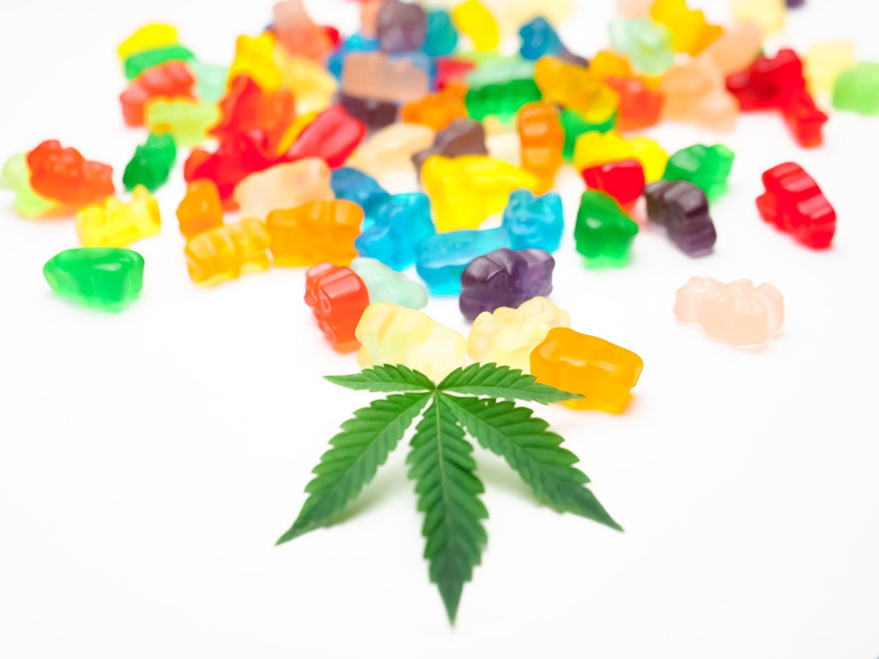 Vibrant gummy bears scattered alongside a cannabis leaf on a white background.